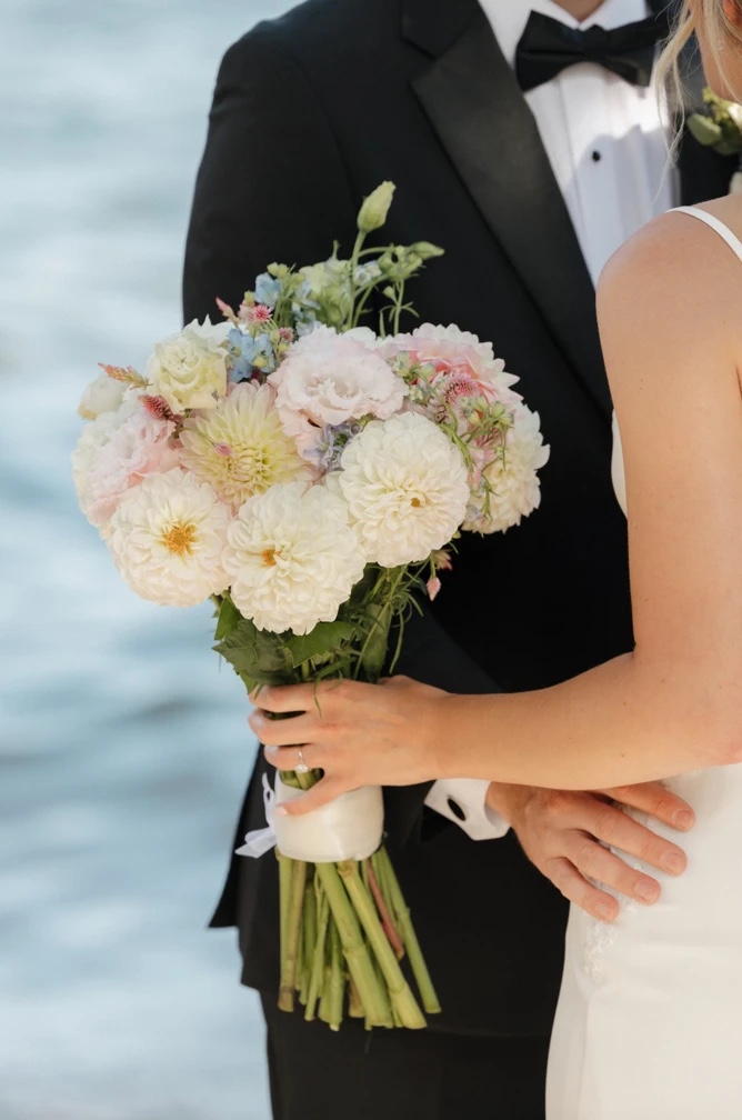 The bridal bouquet of white dahlias and soft pastel blooms, held between the couple by the water