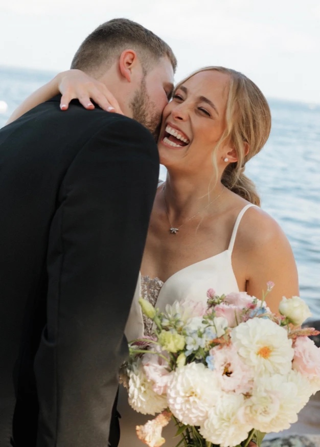 The bride laughing as the groom kisses her cheek at the water's edge, her bouquet in hand
