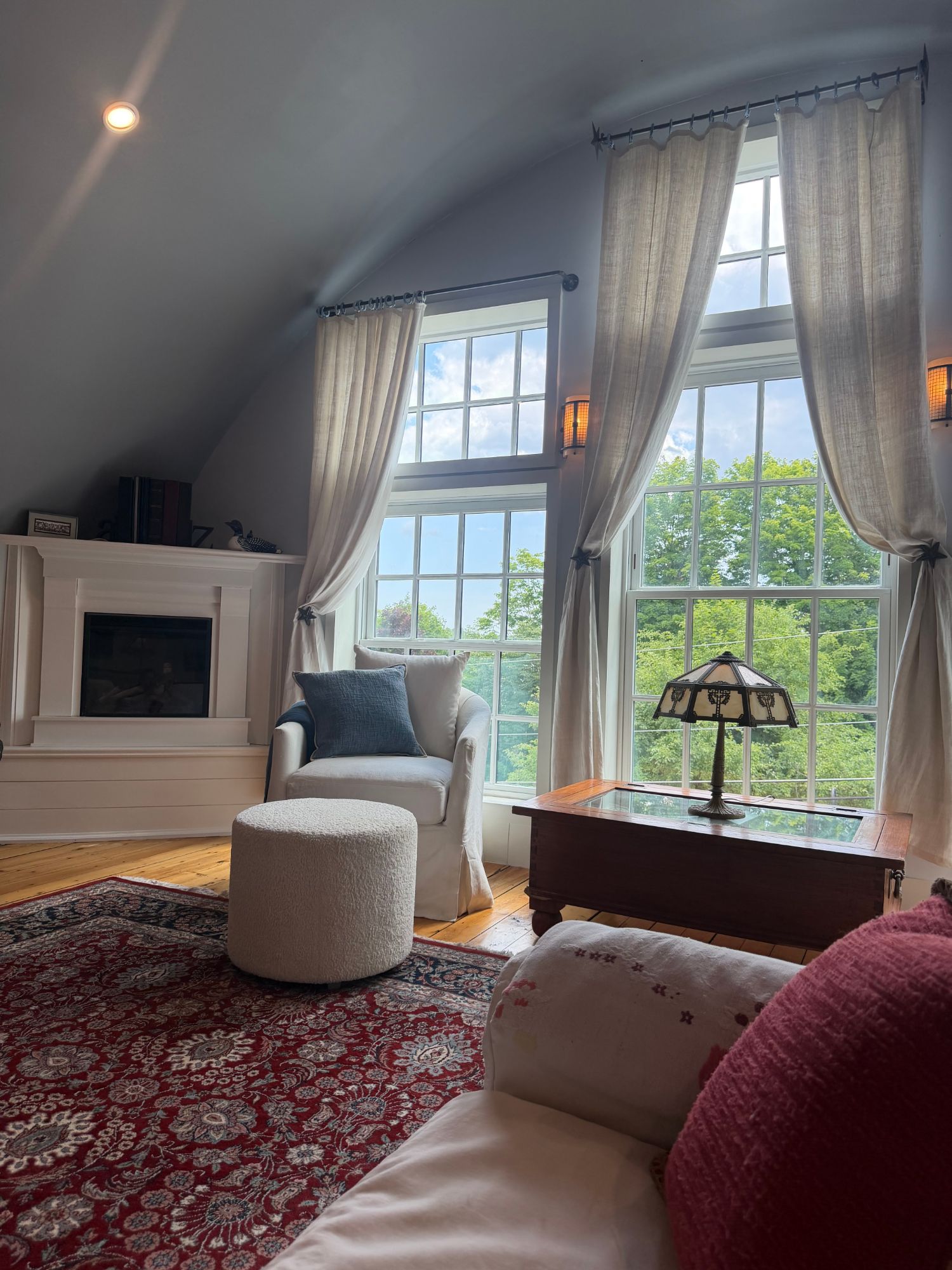 The primary suite's reading nook — a linen chair and fireplace by tall windows overlooking the trees